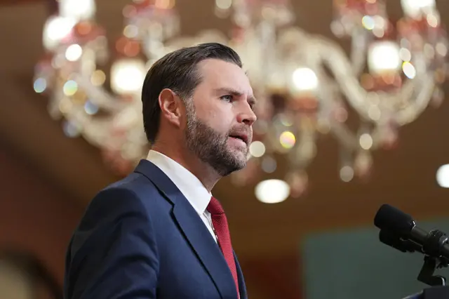 JD Vance, wearing a blue suit and red tie, stands before a microphone with a large chandelier behind him