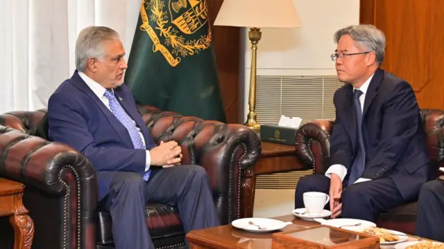 Dar and Zaidong face each other to speak as they sit on separate brown leather sofas next to one another