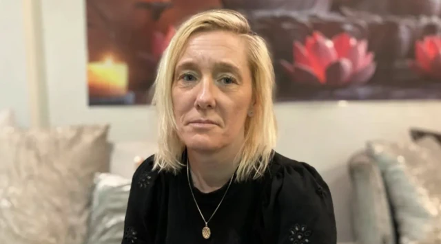 A woman with blonde hair sits on a sofa and looks at the camera