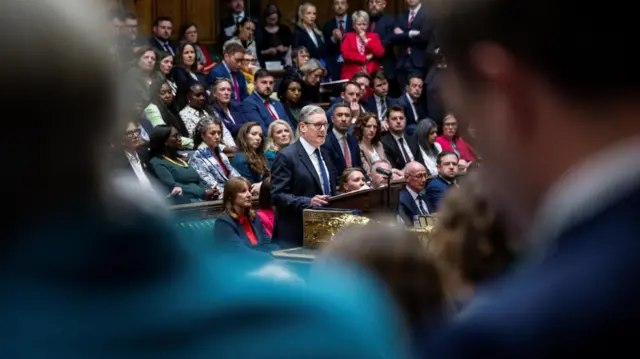 Keir Starmer stands up to give a statement in the House of Commons, with numerous Labour MPs behind him. He is being viewed between the shoulders of two opposition MPs.