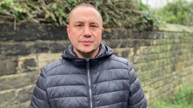 Chris Wild looks at the camera and stands by a wall outside. He wears a black padded jacket.