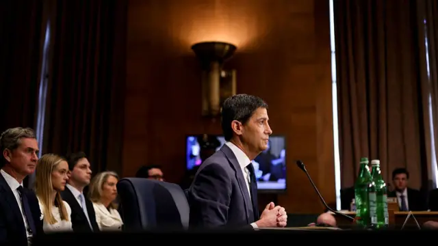 A side view of Kevin Warsh sitting for Senate questioning. Audience members can be see behind him