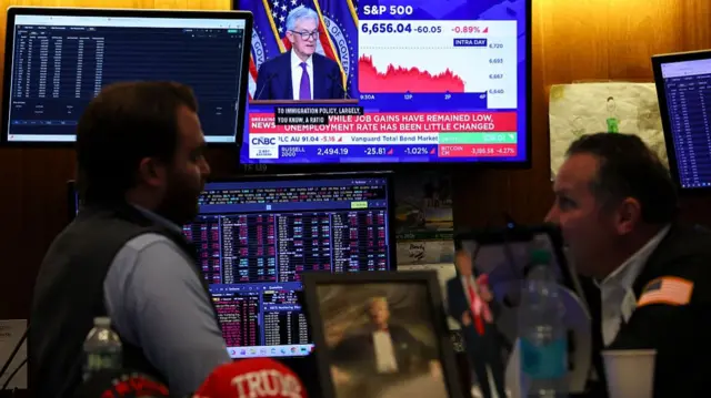 Traders stand in front of a bank of screens, one of them showing Fed Chair Jerome Powell, with Trump memorabilia in the foreground