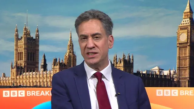 Ed Miliband speaks with the BBC Breakfast backdrop behind him, depicting the Palace of Westminster. He is wearing a navy suit and a dark red tie.