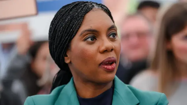 Kemi Badenoch speaking in front of a crowd in a close-up photo. She's looking to the side of the camera and is mid speech. She's wearing a turquoise blazer with a black blouse underneath.