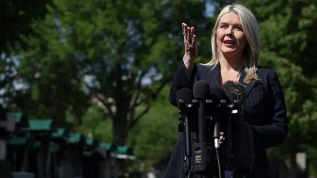 Leavitt, wearing all black, speaking in front of microphones with her hand outstretched in front of green trees