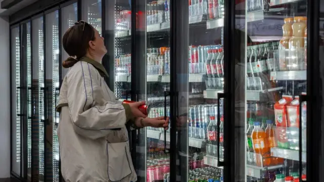 A woman holds a drink and stares at other cans in a fridge in a super market