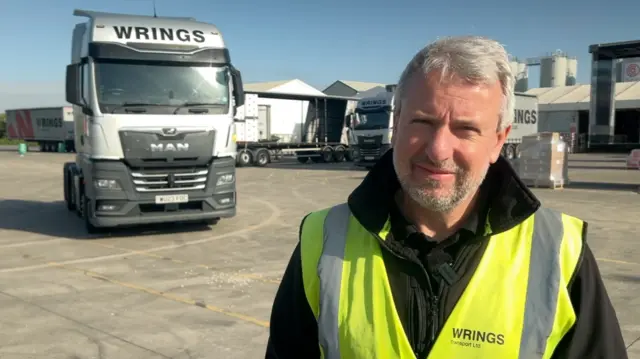 A man stood in a yard with a large white lorry behind him. He is wearing a reflective yellow jacket. He has short grey hair and a short beard.