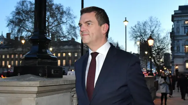 Olly Robbins walks on a busy high street wearing a dark suit and dark red tie
