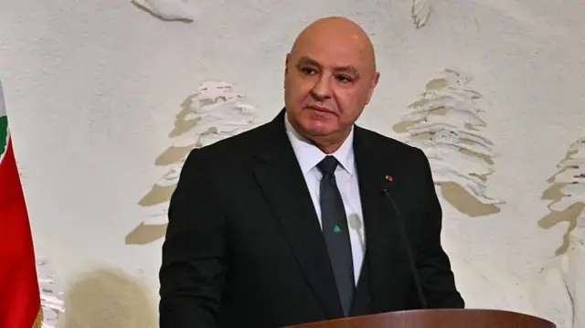 Headshot of Aoun who looks to the side while he stands at a dark wooden podium which has a small microphone on a stand pointing up at him