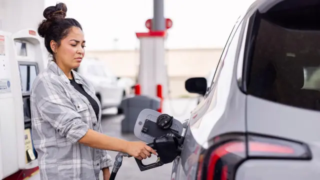 A woman in a grey checked shirt fills up a car at a petrol station.