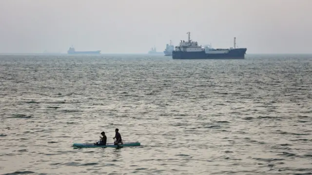 wo boys paddle-board in the sea as ships are anchored near the shoreline on April 22, 2026 in Bandar Abbas, Iran