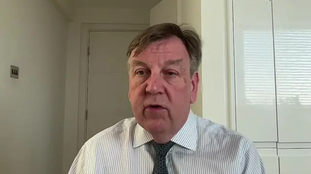 Sir John Whittingdale sits in a room with white walls and a white door. He is wearing a striped shirt and a dark coloured tie.