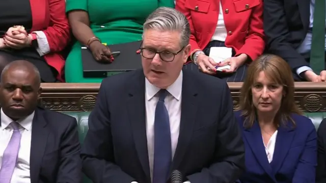 Keir Starmer stands in the House of Commons, with David Lammy and Rachel Reeves sitting on the benches behind him. Starmer is wearing a dark coloured suit and a navy tie