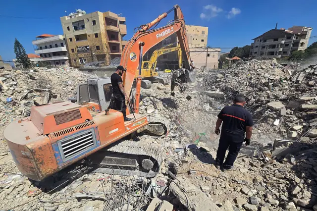 Two diggers operate amid piles of rubble. A man stands on the outside of one of them, and another man is surveying the scene.