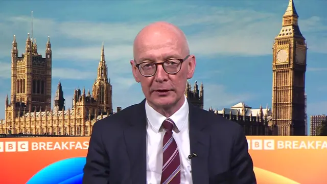 Pat McFadden sits in front of the BBC Breakfast backdrop, which shows the Palace of Westminster. He is wearing a dark coloured suit and a striped red tie.