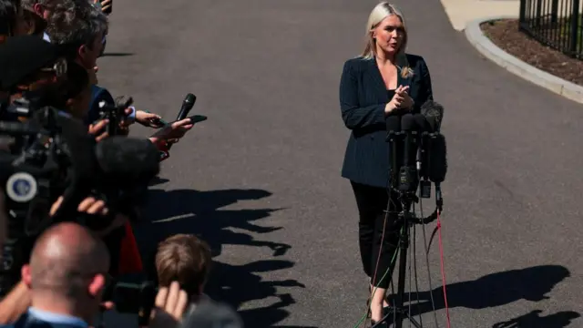 Karole Leavitt standing in front of microphones as reporters with cameras look on