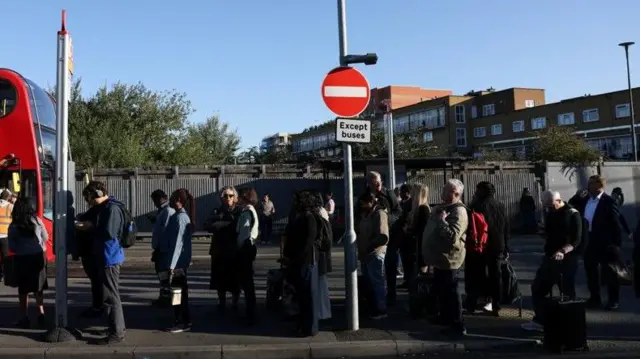 Passengers queue in a line at a bus stop with a red bus just in shot going in the opposite direction. There is a block of flats in the background. Some of the travellers have suitcases with them.