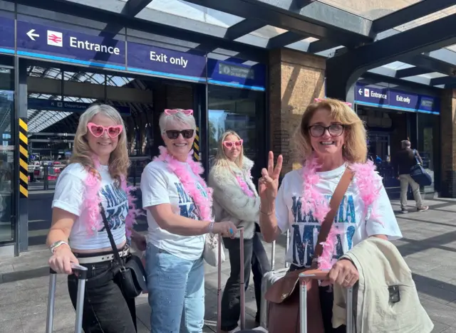 Four women wearing pink sunglasses and feather boas standing outside of King's Cross station