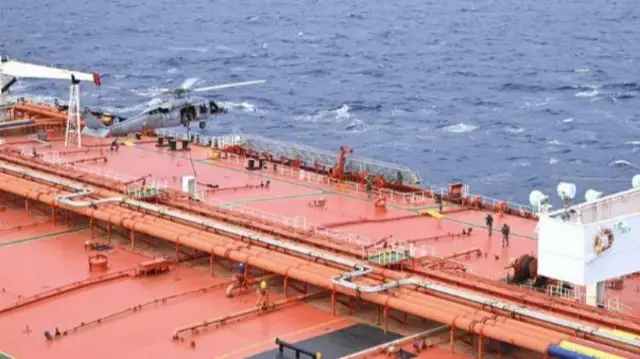 A US military helicopter hovering above the orange deck of a tanker.