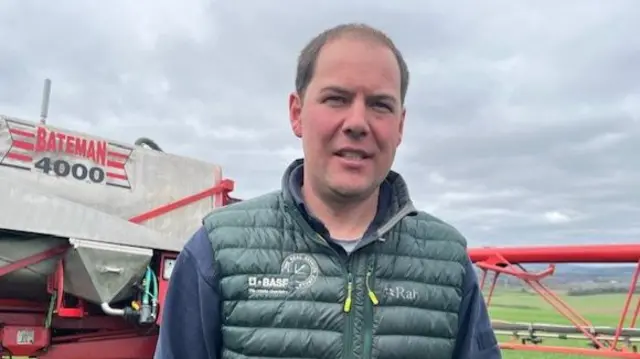 Farmer Ian Campbell standing in a field in front of farm machinery. He is wearing a green quilted body warmer over a blue top