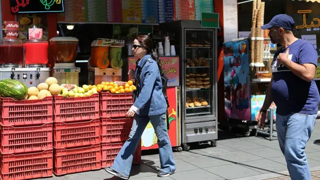 A woman in a blue outfit walks past fresh produce in Tehran
