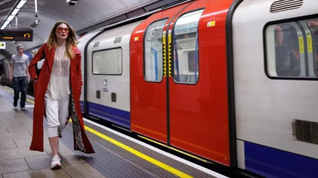 A woman wearing a red coat and orange sunglasses walks next to a Tube train on Thursday