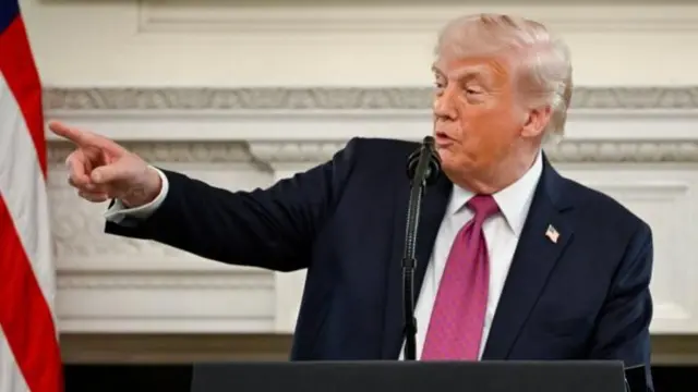 Donald Trump speaks behind a microphone, wearing a blue suit, white shirt and dark pink tie. He is gesturing with his right hand, pointing to his side.