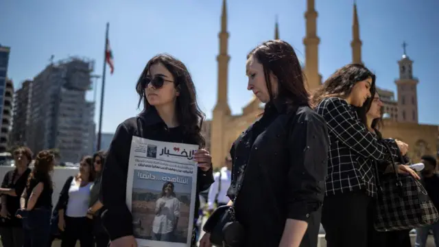 Two women, one holding a newspaper with Amal Khalil's image on the front page, stand in a public square while wearing black.