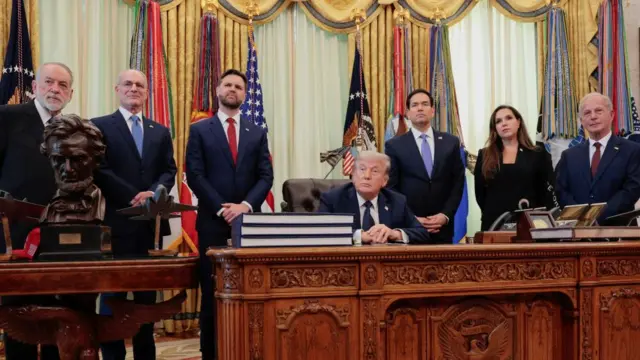 Donald Trump sits at his Oval Office desk, flanked by the vice president, secretary of state and four ambassadors