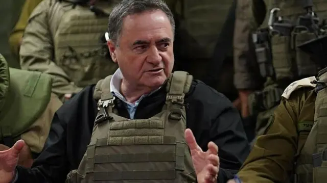 Israel Katz is pictured wearing a khaki military vest and dark jacket. He is gesturing with his hands and looking off-camera.
