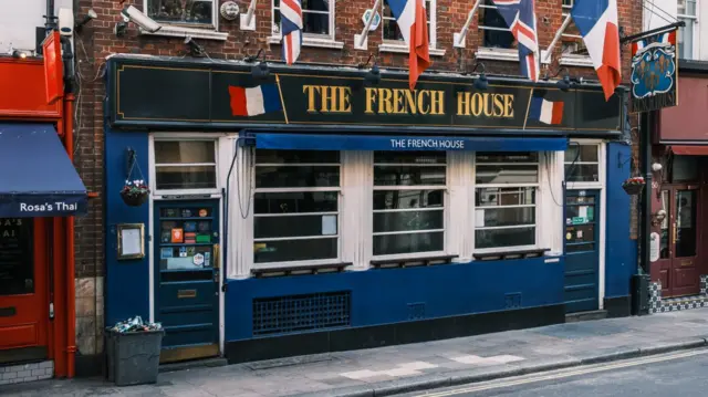 The blue front of The French House can be seen with the door closed. Several French and Union Flags can be seen hanging above the bar
