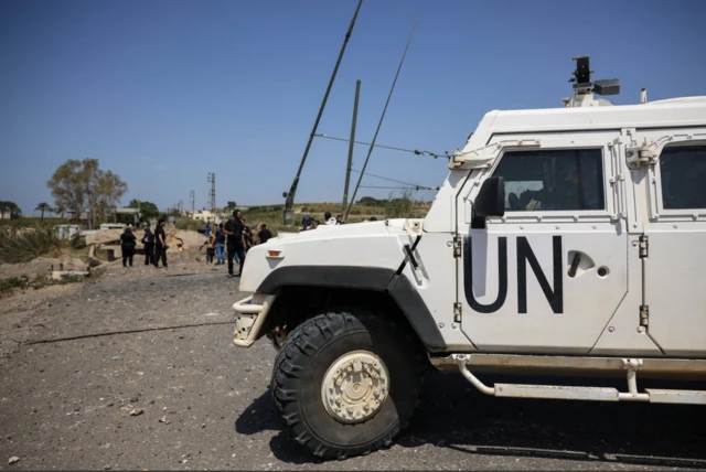 A white UN patrol car parked on a road in Lebanon, a group of people walking by in the background