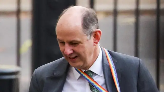 Philip Barton walks in Downing Street. He wears a grey suit, white shirt and green tie. A rainbow patterned lanyard hangs round his neck