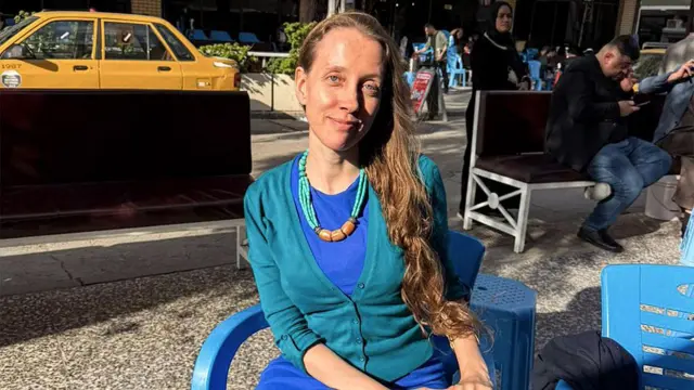 Shelly Kittleson, who has long dark blonde hair and wears a blue dress with a turquiose cardigan, sits on a plastic chair in the sun in a Baghdad street