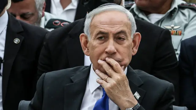 Benjamin Netanyahu in dark suit, white shirt and blue tie sits on a chair, his left hand resting on the lower part of his face