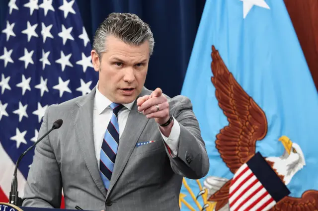 Pete Hegseth in a grey suit pointing in front of him with his left hand. Behind him as US flag and a DoD flag