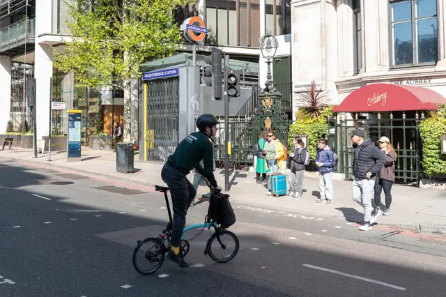 A person on a foldable bike cycles on a road as people stand on a pavement beside Knightsbridge station which has its entrance shutters closed