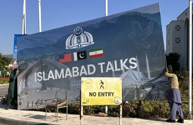 Two men carry a sign with the wording Islamabad talks to place it in patch of grass. Another sign, in yellow metal, is in the foreground; it reads "no entry"