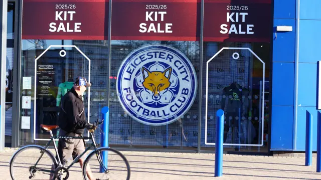 A photograph of Leicester City's clubshop advertising a sale of their replica kit with a man with a bicycle walking past