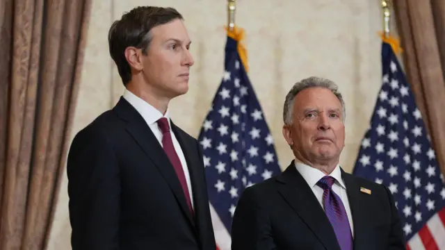 Jared Kushner, with short brown hair, dressed in a suit and tie, stands next to Steve Witkoff, a shorter man with grey hair also dressed in a suit. Two US flags are placed behind them
