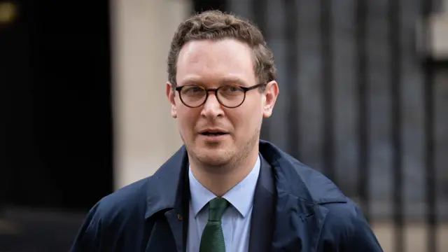 Darren Jones a man with brown wavy hair and glasses, dressed in a blue suit and overcoat