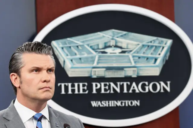 Pete Hegseth stands to the left of the frame, a wooden panel bearing a drawing of the Pentagon building behind him