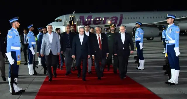 A group of men in suits walks towards the camera away from a grey passenger plane on a red carpet flanked by military personnel in light blue uniforms