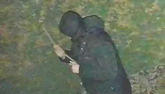 A still from a video showing John Ashby with a long stick, wearing a dark coat with his hood up