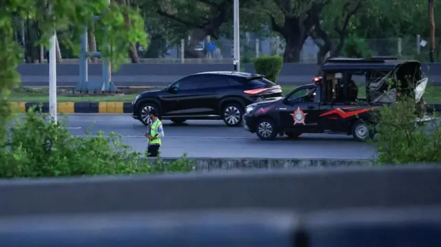 A black car flanked by another police vehicle drives down a road