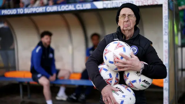 A member of the Rochdale staff clasps four balls as he stands by the dugout