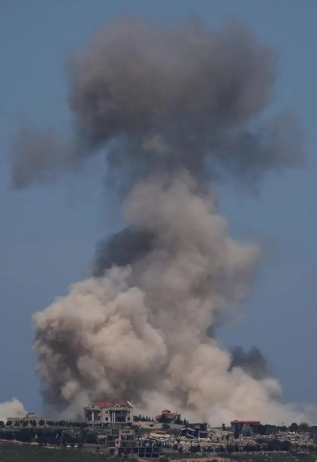 Thick grey smoke rises in the sky over a green plain with several houses visible