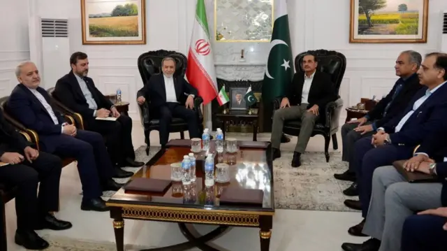 Several men sit at chairs around a table with the Iranian and Pakistani flags in the background