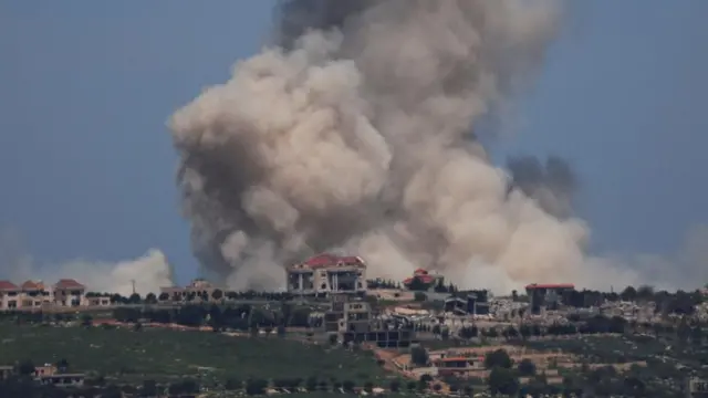 Thick grey smoke rises in the sky over a green plain with several houses visible